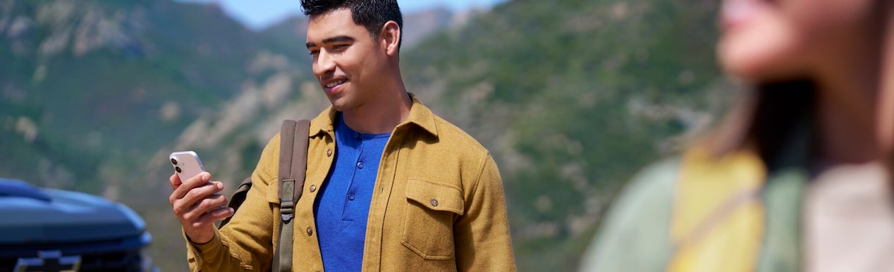 A smiling man with a backpack stands in a rugged, mountainous landscape, looking down at his smartphone, with a vehicle partially visible behind him.