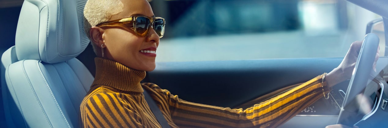 A woman with sunglasses on is smiling with her left hand on the steering wheel