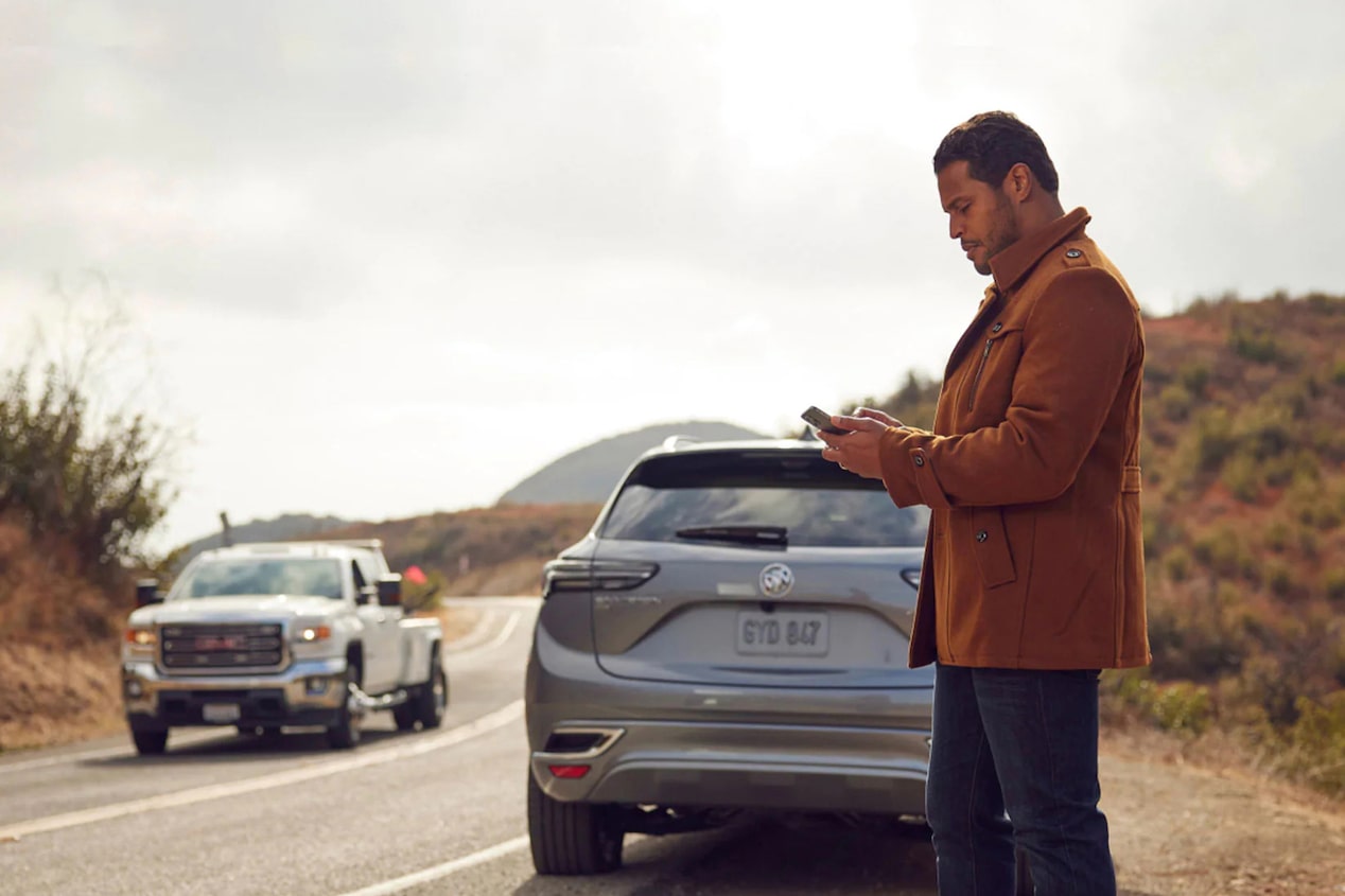 Man Standing at the Rear of Buick Looking at His Phone as a white truck drives on the opposite side of the road