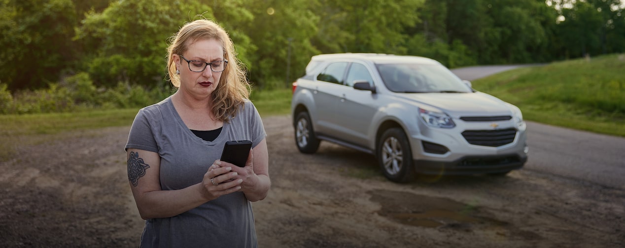 Nicole Kutas looks down at her phone while standing next to her Chevrolet vehicle that is parked on a dirt road.