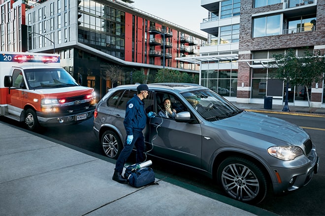 A First Responder talks to someone in the passenger seat of a vehicle parked at a curb, with an ambulance parked behind it.