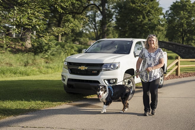 A woman walks her dog near her Chevy vehicle that is parked on the side of a road.