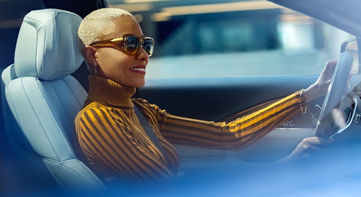 A woman in a mustard yellow turtleneck and sunglasses is in the driver seat of a vehicle with her left hand on the steering wheel.